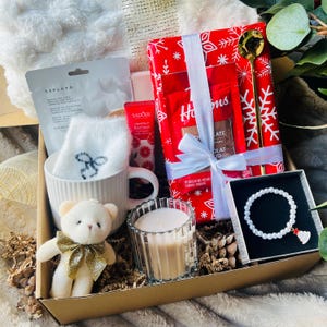 May include: A gift box containing a white mug with fuzzy socks, a small plush bear with a gold bow, a glass candle, a pearl bracelet with a Christmas tree charm, a gold spoon, a red gift-wrapped package, hand cream, and a facial sheet mask.
