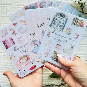 May include: A set of rectangular notebooks with light blue and white vertical stripes. The covers feature illustrations of books, teacups, armchairs, and windows. The text on the covers reads, "Live. Love. Read." and "This is your day. Enjoy it!"