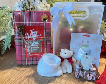 Op de afbeelding: Een cadeaupakket met een rood geruite doos, een doorzichtige tas met een witte strik en een kleine witte teddybeer met een rode sjaal. Inclusief een Tim Hortons warme chocolademelk zakje en een gouden lepel.
