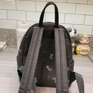 May include: A dark gray backpack with black straps and handle. The backpack features a repeating pattern of green and white cartoon characters and the letters "NYC". The backpack has two adjustable shoulder straps.