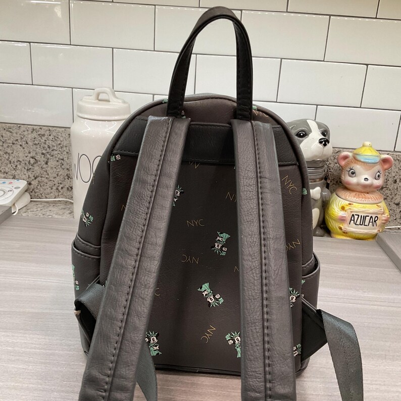 May include: A dark gray backpack with black straps and handle. The backpack features a repeating pattern of green and white cartoon characters and the letters "NYC". The backpack has two adjustable shoulder straps.
