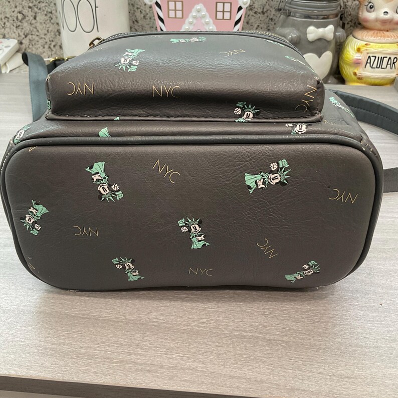 May include: A dark gray backpack with a small front pocket. The bag is patterned with small green and white Statue of Liberty graphics and the letters "NYC" in gold. The bag has a top handle and a zipper closure.