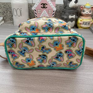 May include: A beige backpack featuring a repeating pattern of the character Stitch in various poses and outfits. The backpack has a teal green trim and a top handle. The design includes Stitch in blue, orange, and pink.