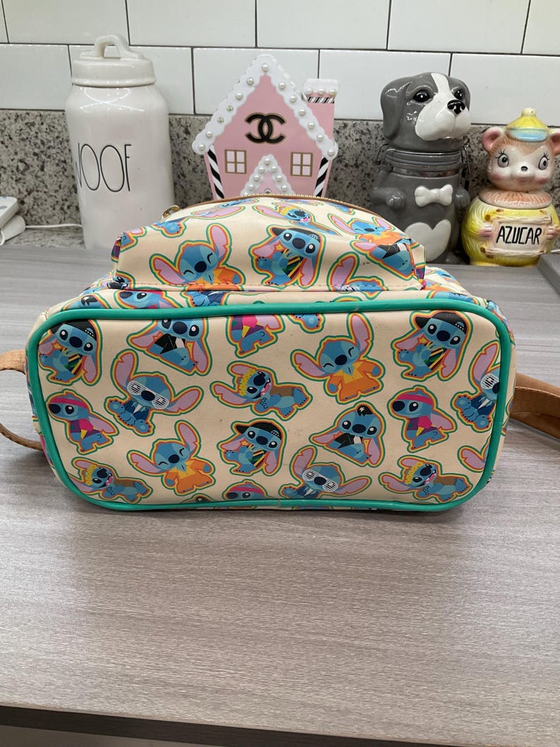 May include: A beige backpack featuring a repeating pattern of the character Stitch in various poses and outfits. The backpack has a teal green trim and a top handle. The design includes Stitch in blue, orange, and pink.