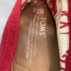 May include: Close-up of a red TOMS shoe interior. The shoe's insole is brown, with the brand name and size W7.5 visible. The inner lining features a cream-coloured fabric with a repeating red pattern. The text reads: "With every pair you purchase, TOMS will give a pair of new shoes to a child in need. One for One."