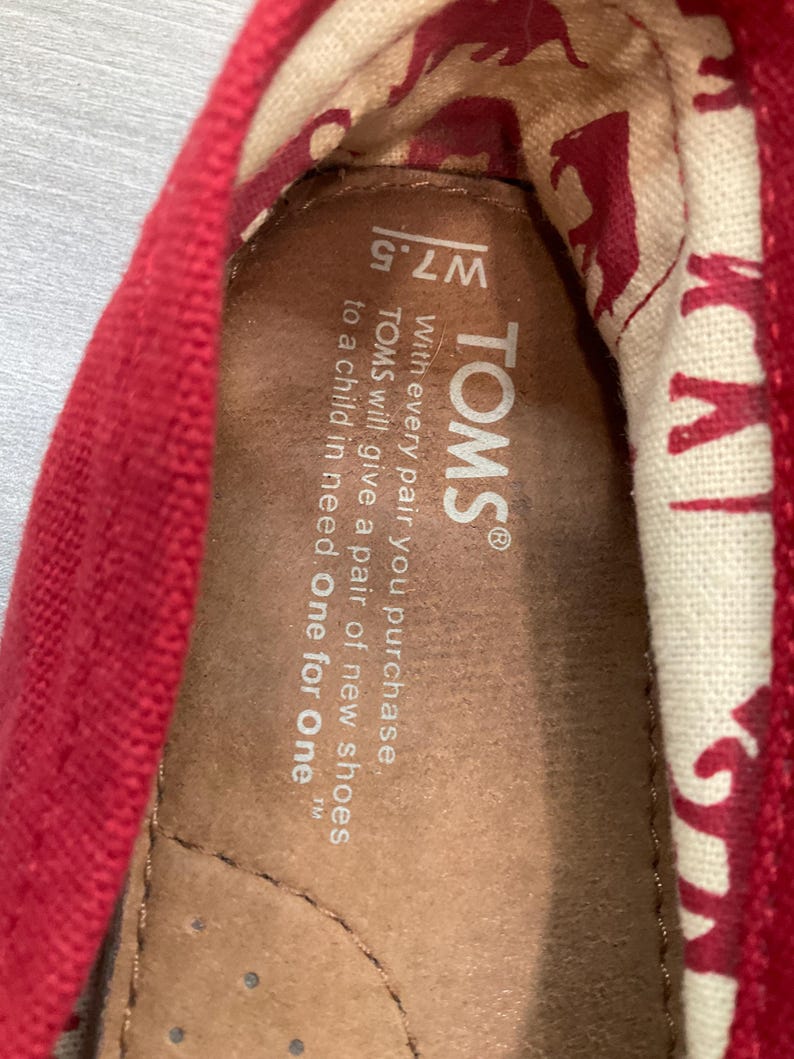 May include: Close-up of a red TOMS shoe interior. The shoe's insole is brown, with the brand name and size W7.5 visible. The inner lining features a cream-coloured fabric with a repeating red pattern. The text reads: "With every pair you purchase, TOMS will give a pair of new shoes to a child in need. One for One."