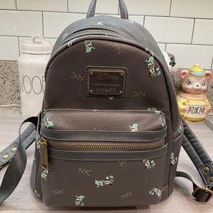 May include: Gray faux leather backpack with a front pocket and top handle. The backpack features a repeating pattern of green floral designs and the letters "NYC". A rectangular plaque reads "Disney Loungefly".