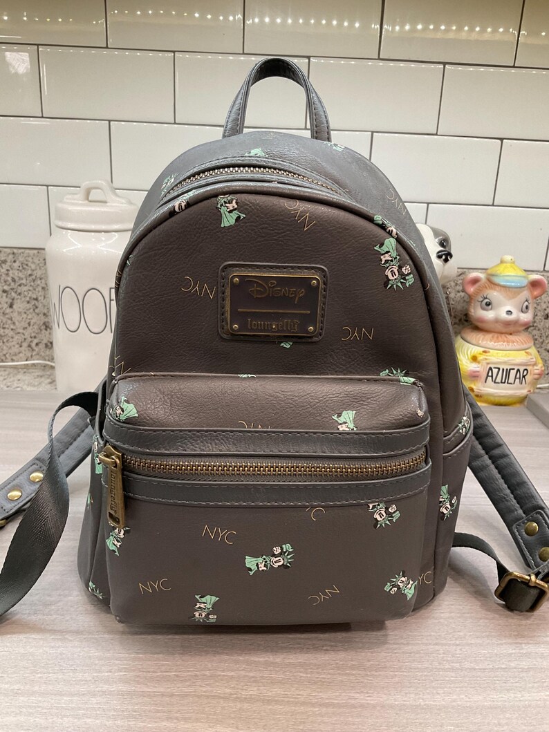 May include: Gray faux leather backpack with a front pocket and top handle. The backpack features a repeating pattern of green floral designs and the letters "NYC". A rectangular plaque reads "Disney Loungefly".