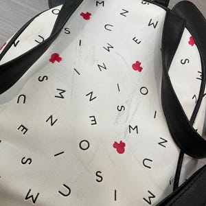 May include: White backpack with black straps and a Mickey Mouse ear design. The bag features a pattern of black letters and red Mickey Mouse head shapes. The straps and ears are black, and the bag has a silver buckle.