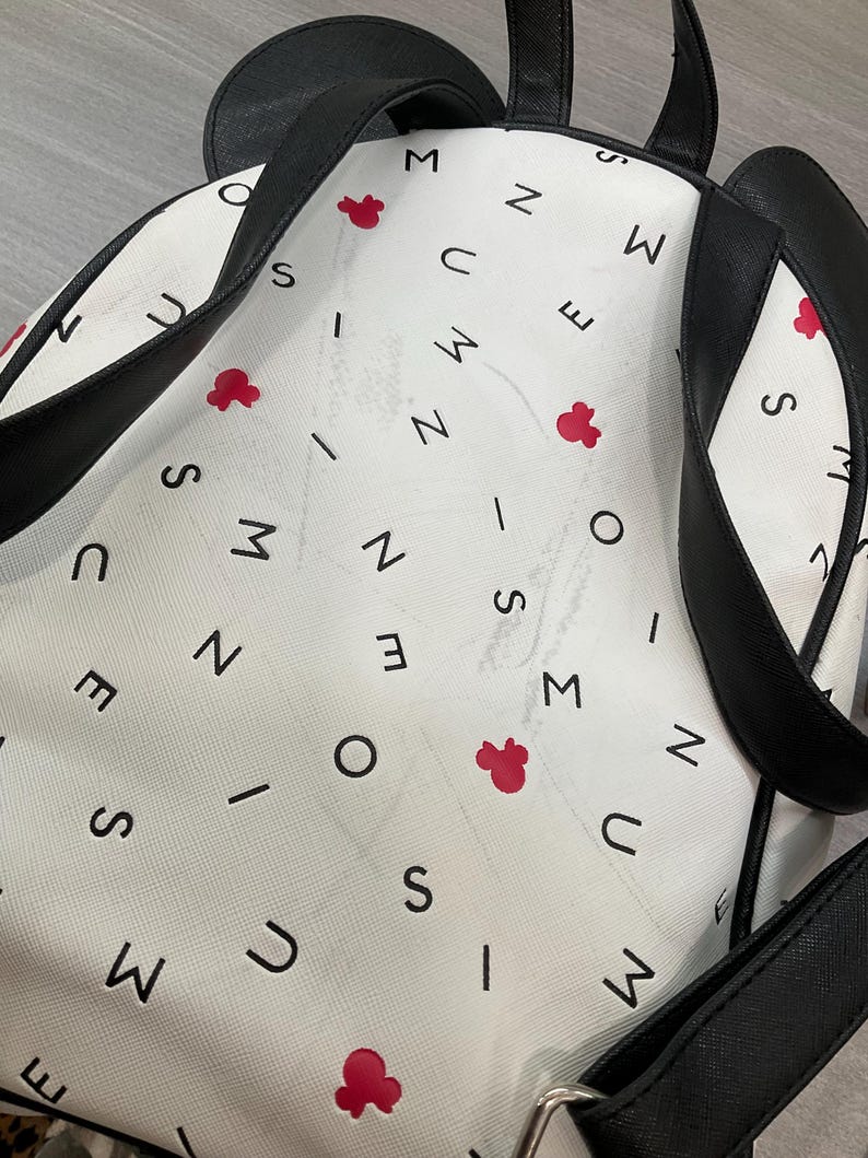 May include: White backpack with black straps and a Mickey Mouse ear design. The bag features a pattern of black letters and red Mickey Mouse head shapes. The straps and ears are black, and the bag has a silver buckle.