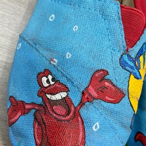 May include: Close-up of blue canvas shoes with red trim, hand-painted with a cartoon crab and other sea creatures. The shoes feature the brand name "TOMS" on the red trim. The shoes are a size 7 UK.