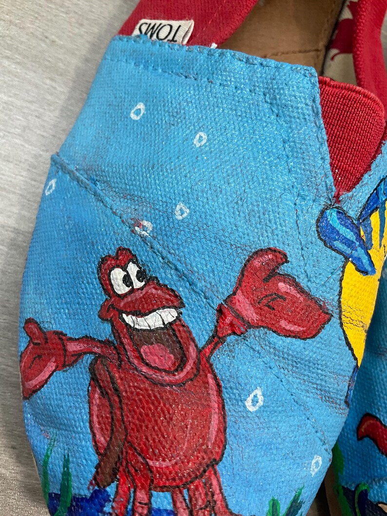 May include: Close-up of blue canvas shoes with red trim, hand-painted with a cartoon crab and other sea creatures. The shoes feature the brand name "TOMS" on the red trim. The shoes are a size 7 UK.
