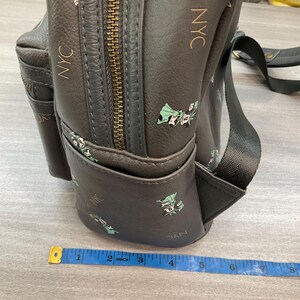 May include: Dark gray backpack with a repeating pattern of the Statue of Liberty and the letters "NYC" in gold. The backpack has a zippered main compartment and a small front pocket. A measuring tape is in the foreground.
