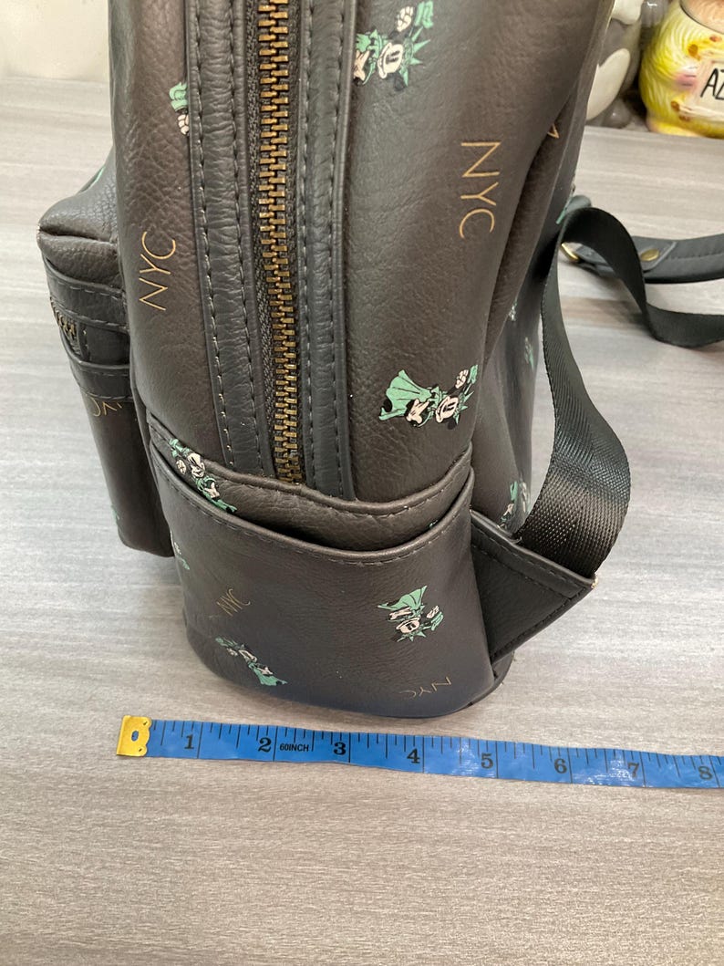 May include: Dark gray backpack with a repeating pattern of the Statue of Liberty and the letters "NYC" in gold. The backpack has a zippered main compartment and a small front pocket. A measuring tape is in the foreground.