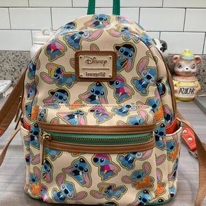 May include: A beige Disney Loungefly mini backpack featuring a repeating pattern of the character Stitch in various outfits. The backpack has brown trim, a green handle, and a front pocket with a gold zipper. The Disney Loungefly logo is on a brown patch.