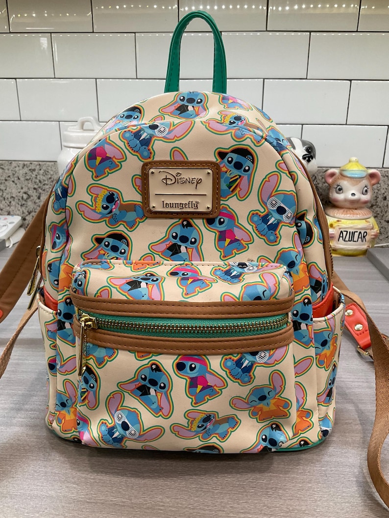 May include: A beige Disney Loungefly mini backpack featuring a repeating pattern of the character Stitch in various outfits. The backpack has brown trim, a green handle, and a front pocket with a gold zipper. The Disney Loungefly logo is on a brown patch.