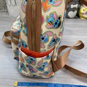 May include: A beige backpack featuring a repeating pattern of the character Stitch in various poses and colors. The backpack has brown trim, a front pocket with an orange interior, and a gold zipper. The straps are a tan color.