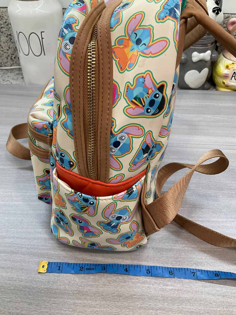 May include: A beige backpack featuring a repeating pattern of the character Stitch in various poses and colors. The backpack has brown trim, a front pocket with an orange interior, and a gold zipper. The straps are a tan color.