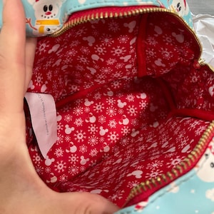 以下が含まれることがあります： 赤色の内装の、クリスマスをテーマにした水色のバックパック。外側には漫画の雪だるま、キャンディケイン、靴下が描かれています。内側の裏地は赤色で、白い雪の結晶とミッキーマウスのシルエットが描かれています。バッグには金のジッパーが付いています。