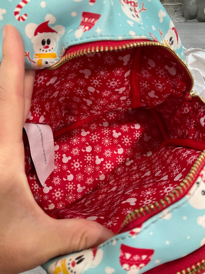 以下が含まれることがあります： 赤色の内装の、クリスマスをテーマにした水色のバックパック。外側には漫画の雪だるま、キャンディケイン、靴下が描かれています。内側の裏地は赤色で、白い雪の結晶とミッキーマウスのシルエットが描かれています。バッグには金のジッパーが付いています。