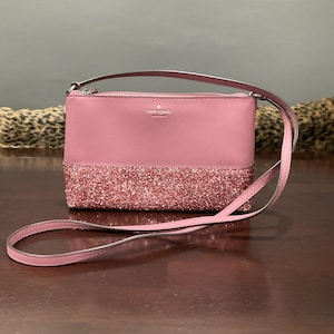 Puede incluir: Un bolso cruzado rosa empolvado con una parte inferior brillante y una parte superior lisa. El bolso tiene una correa fina a juego y un cierre de cremallera. La marca "kate spade" es visible en la parte delantera.