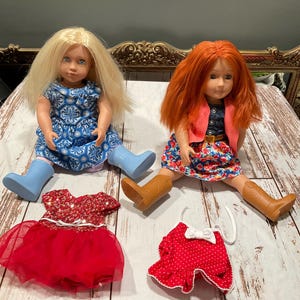 May include: Two dolls with blonde and red hair, wearing dresses and boots. One doll is in a blue floral dress and blue boots, the other in a floral dress, a pink jacket, and brown boots. Two additional doll outfits are also present.