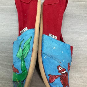 May include: Pair of TOMS slip-on shoes. The shoes have red canvas uppers and blue canvas toes with an underwater scene featuring a green plant and a red crab. The soles are beige.