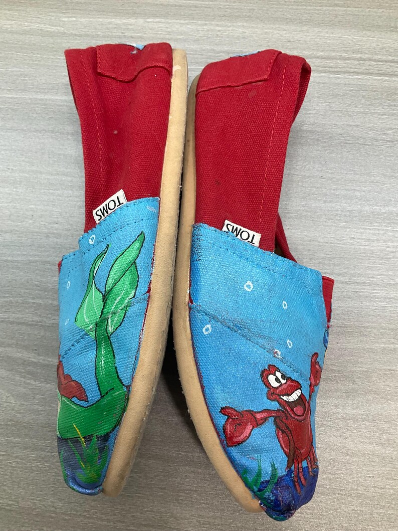 May include: Pair of TOMS slip-on shoes. The shoes have red canvas uppers and blue canvas toes with an underwater scene featuring a green plant and a red crab. The soles are beige.