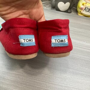 May include: A pair of red canvas slip-on shoes with a light brown sole. Each heel displays a blue and white label with the brand name "TOMS".