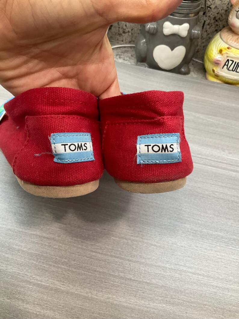 May include: A pair of red canvas slip-on shoes with a light brown sole. Each heel displays a blue and white label with the brand name "TOMS".