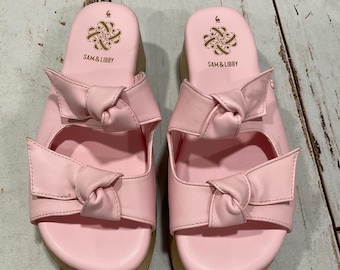 May include: A pair of light pink sandals with bow accents. The sandals have a slip-on design with a rounded toe and a small platform sole. The brand name "SAM & LIBBY" is printed on the footbed. The sandals are size 6.