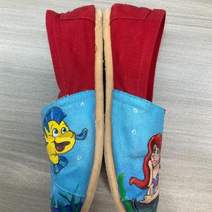 May include: Canvas slip-on shoes with red uppers and light blue toes. One toe features a yellow cartoon fish, the other a red-haired mermaid. The shoes have beige soles. The shoes are decorated with cartoon characters.