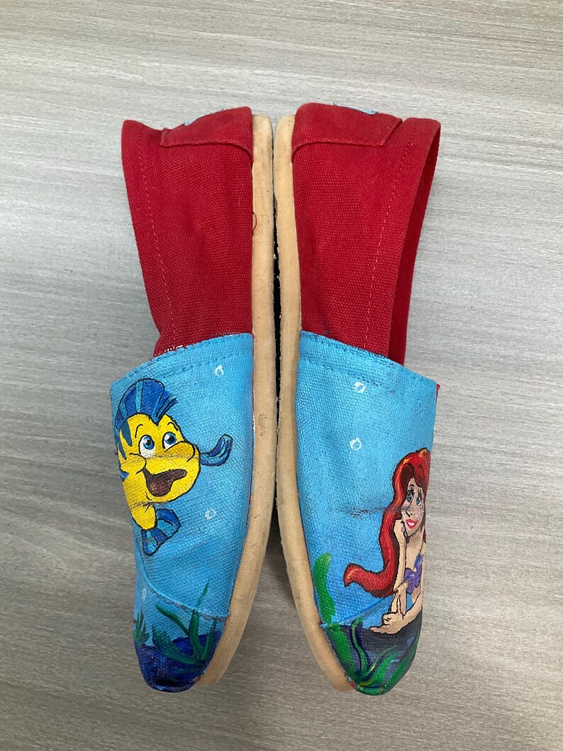 May include: Canvas slip-on shoes with red uppers and light blue toes. One toe features a yellow cartoon fish, the other a red-haired mermaid. The shoes have beige soles. The shoes are decorated with cartoon characters.