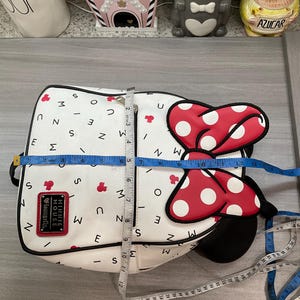 May include: White backpack with a Minnie Mouse design. The bag features a large red bow with white polka dots and a black outline. The bag is approximately 12 inches wide and 12 inches tall. The bag has the text "MINNIE MOUSE Loungefly".