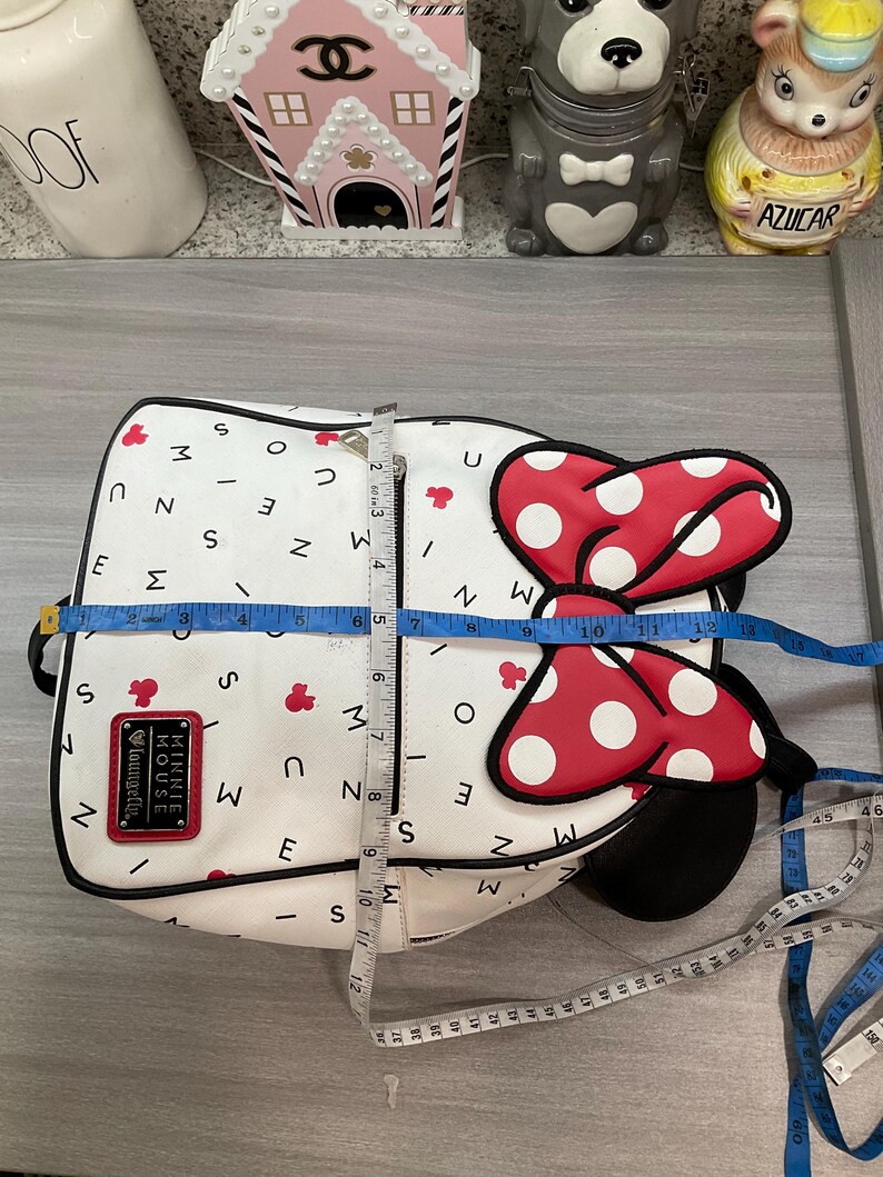May include: White backpack with a Minnie Mouse design. The bag features a large red bow with white polka dots and a black outline. The bag is approximately 12 inches wide and 12 inches tall. The bag has the text "MINNIE MOUSE Loungefly".