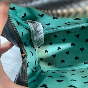 May include: Black zippered handbag with a teal interior featuring black Mickey Mouse silhouettes. The interior shows a white tag with printed text. The bag's zipper is gold-colored.