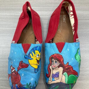 May include: Pair of TOMS slip-on shoes with a red canvas upper and a blue painted design. The design features characters from The Little Mermaid: Ariel, Flounder, and Sebastian. The shoes are size W7.
