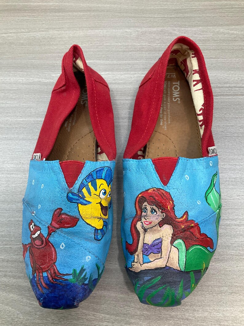 May include: Pair of TOMS slip-on shoes with a red canvas upper and a blue painted design. The design features characters from The Little Mermaid: Ariel, Flounder, and Sebastian. The shoes are size W7.