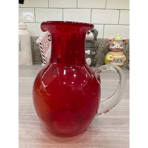 Puede incluir: Una jarra de cristal de color rojo vibrante con cuerpo redondeado, borde ancho y acampanado, y un asa de cristal transparente unida al lateral. La jarra tiene una superficie lisa y translúcida que capta la luz, mostrando su color carmesí.