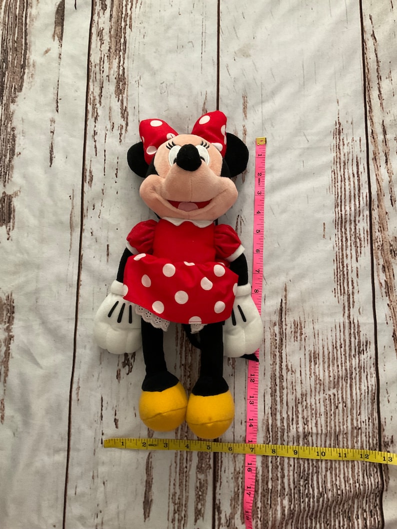 May include: A Minnie Mouse plush doll, featuring a red dress with white polka dots and a matching bow. The doll has black ears, a pink face, and yellow shoes. The doll is approximately 13 inches tall.