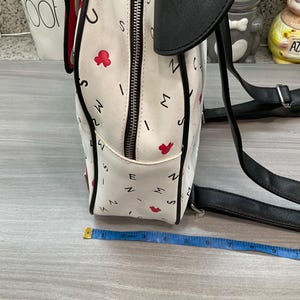 May include: White backpack with black accents, featuring a pattern of letters and red flower shapes. The backpack has a black top flap and straps. A measuring tape is placed near the bag.