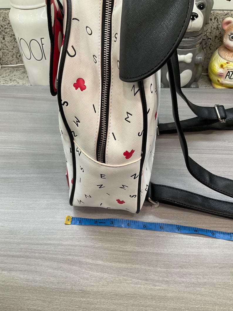 May include: White backpack with black accents, featuring a pattern of letters and red flower shapes. The backpack has a black top flap and straps. A measuring tape is placed near the bag.
