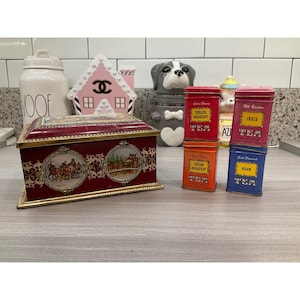 May include: A rectangular maroon metal tea tin with gold trim and two circular illustrations of horse-drawn carriages, accompanied by four smaller vertical tea tins in red, orange, pink, and blue, each labelled with tea varieties and net weights.