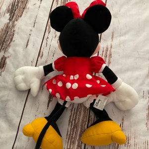 May include: A plush Minnie Mouse doll, viewed from the back, with a red dress with white polka dots, a matching bow, and yellow shoes. The doll has black ears, arms, and legs, and white gloves. The Disney tag is visible.