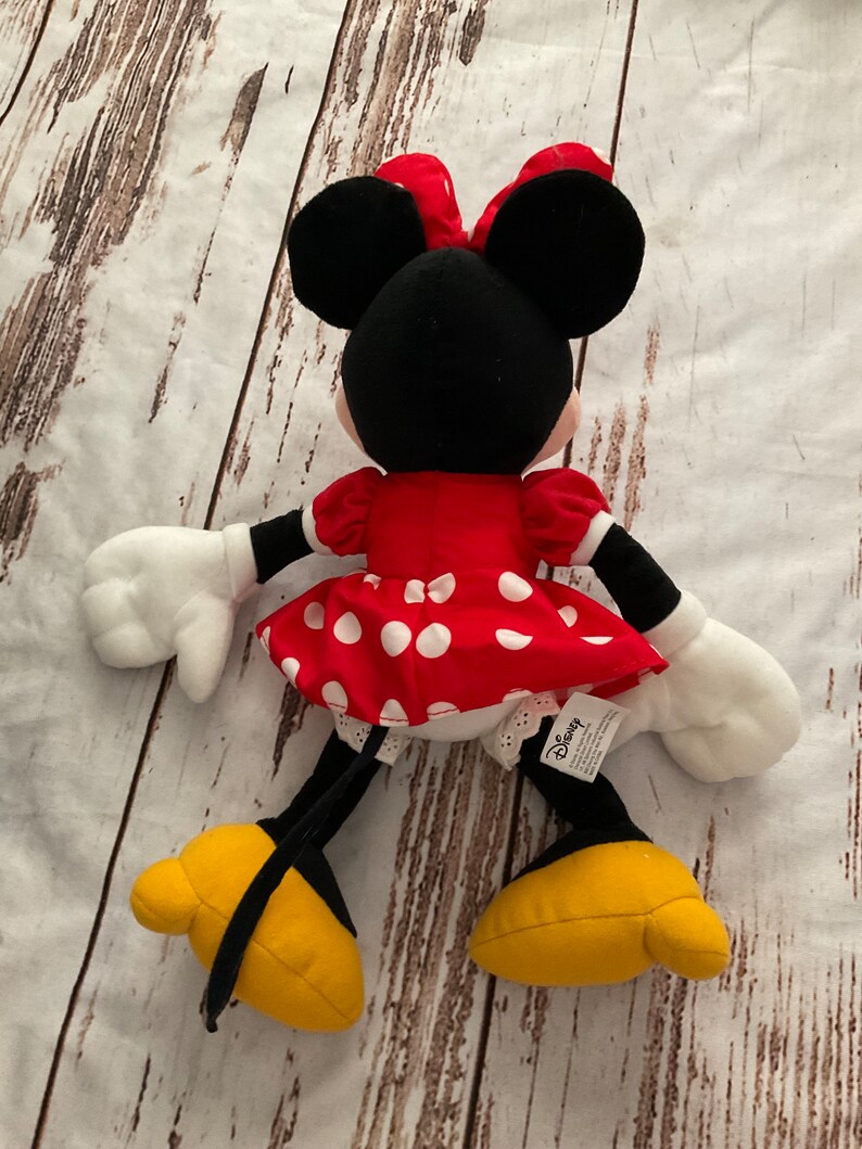 May include: A plush Minnie Mouse doll, viewed from the back, with a red dress with white polka dots, a matching bow, and yellow shoes. The doll has black ears, arms, and legs, and white gloves. The Disney tag is visible.