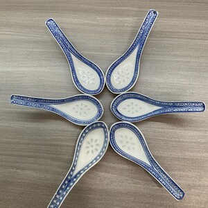 May include: Six white ceramic soup spoons with blue floral and geometric patterns. The spoons are arranged in a circular pattern, showcasing their decorative design. Each spoon has a small hole at the end of the handle.