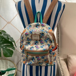 May include: A beige backpack featuring a repeating pattern of the Disney character Stitch in various poses and colors. The backpack has brown straps, a green top handle, and a front pocket with a gold zipper. The Disney and Loungefly logos are visible.