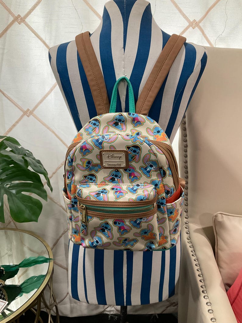 May include: A beige backpack featuring a repeating pattern of the Disney character Stitch in various poses and colors. The backpack has brown straps, a green top handle, and a front pocket with a gold zipper. The Disney and Loungefly logos are visible.