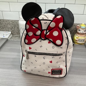 May include: White backpack featuring a red polka dot bow and black mouse ears. The bag has a front zipper pocket and is decorated with red Minnie Mouse icons and the text "MINNIE MOUSE" and "Loungefly".