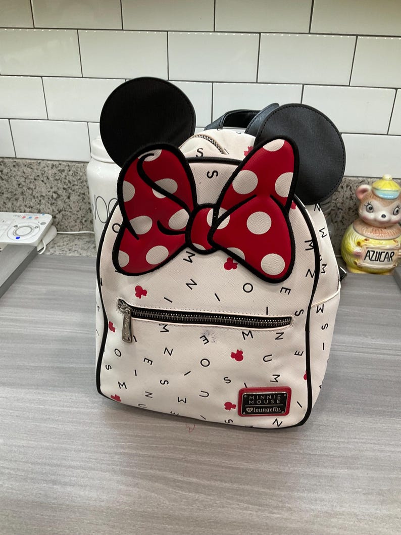 May include: White backpack featuring a red polka dot bow and black mouse ears. The bag has a front zipper pocket and is decorated with red Minnie Mouse icons and the text "MINNIE MOUSE" and "Loungefly".